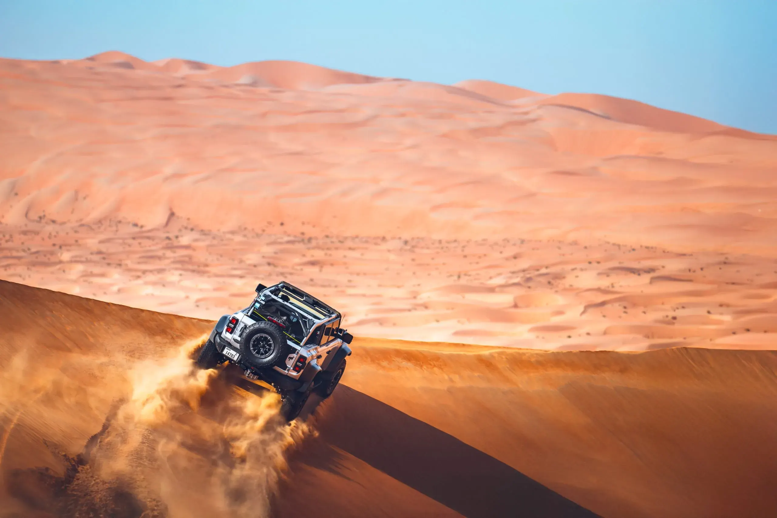 A silver 4x4 cresting a dune ridge under a clear blue sky, sand flying behind it